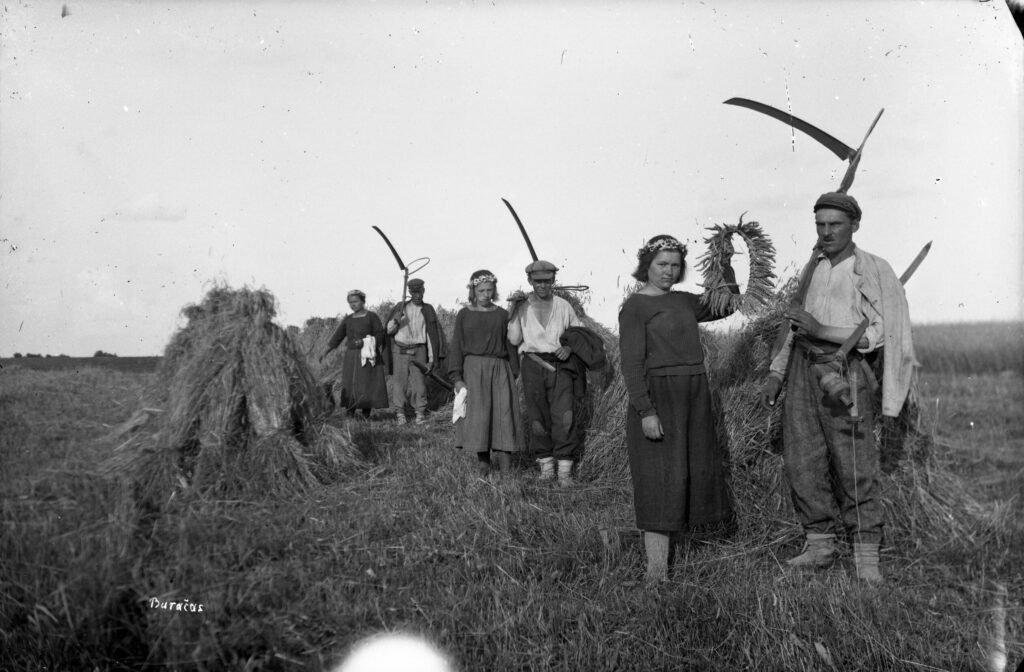 Rugių lauku einantys pjovėjai su dalgiais ir mergina, nešanti pabaigtuvių vainiką